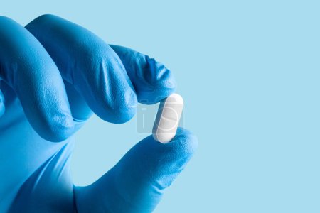Blue-gloved hand holding a white pill against a light blue background