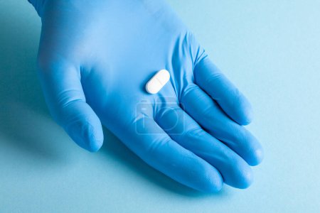 A single white pill placed in a blue-gloved hand against a soft blue background
