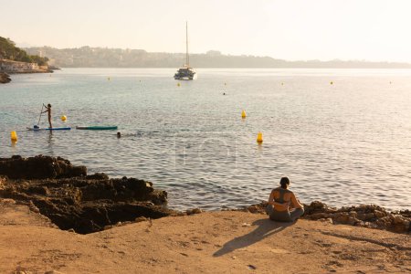 Téléchargez les photos : Ses Illetes, Majorque, Îles Baléares, Espagne. 22 juillet 2022 - Catamaran et personnes faisant du paddle surf près de la côte rocheuse de Cala Comtesa cove, avec une femme faisant du yoga, au lever du soleil - en image libre de droit