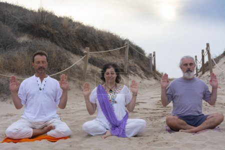 Photo for Group of men and women practicing kundalini yoga on the sand at the beach. Abhaya pose - Royalty Free Image