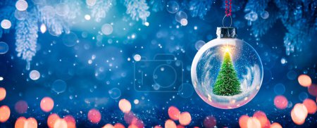 Photo for Glass Ornament With Tree Inside Hanging From Pine Branch And Defocused Lights - Christmas - Royalty Free Image