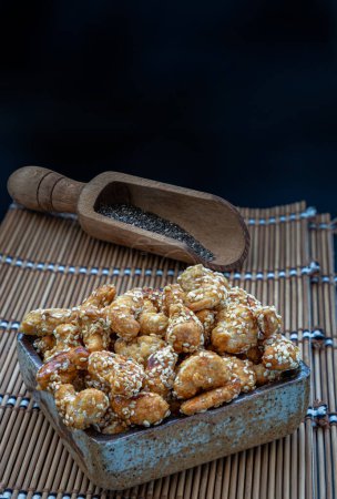 Sweet Cashew Nuts coated with white sesame seeds in the ceramic bowl served with black sesame on bamboo mat. Healthy snack, Space for text, Selective Focus.