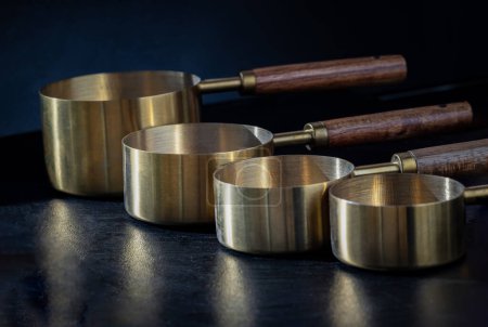 Set of Brass Measuring Cups with Wood Handles with Hanging Hole Design on dark background. Wooden & Brass Measuring Cups & Spoons for Measuring Dry and Liquid Ingredients, Kitchen tool, Space for text, Selective Focus.