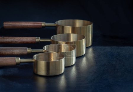 Set of Brass Measuring Cups with Wood Handles with Hanging Hole Design on dark background. Wooden & Brass Measuring Cups & Spoons for Measuring Dry and Liquid Ingredients, Kitchen tool, Copy space, Selective Focus.