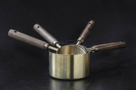 Set of Brass Measuring Cups with Wood Handles with Hanging Hole Design on dark background. Wooden & Brass Measuring Cups & Spoons for Measuring Dry and Liquid Ingredients, Kitchen tool, Space for text, Selective Focus.