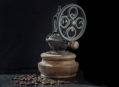 Old original coffee grinder with traces of time and scuffs on the body with coffee beans on dark background. Antique coffee bean original grinder metal shake wheel with hand crank, Copy space, Selective focus.