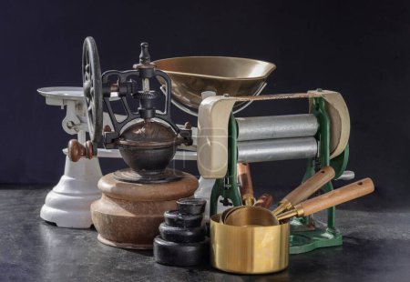 Antique collection has Pasta maker with hand crank, Original coffee grinder metal shake wheel with hand crank, Weight scale and Brass Measuring Cups on Dark background. Space for text. Selective focus.