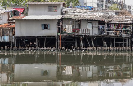 Foto de Bangkok, Tailandia - 20 de abril de 2024 - En la zona de tugurios, las casas son antiguas a lo largo del Khlong Phra Khanong. Casas rotas están al lado del Canal con agua sucia. Problema de contaminación, Copiar espacio, Enfoque selectivo. - Imagen libre de derechos