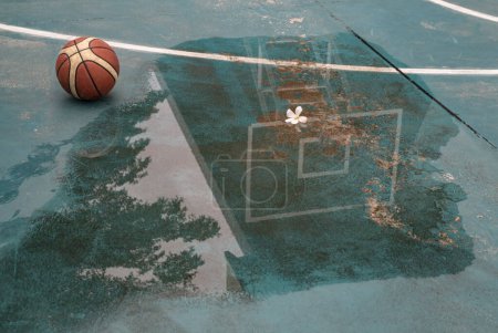 Basketball backboard and hoop with reflection water after rain and Basketball ball on outdoor court. Basketball on wet court, Basketball half-court line, Copy space, Selective focus.