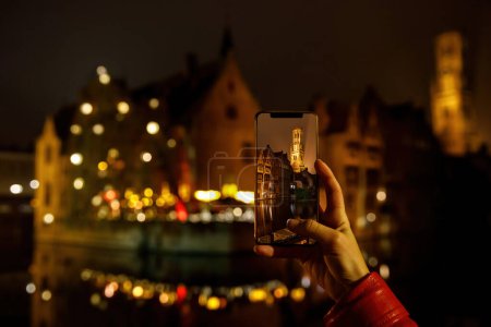 Photo for Taking picture of Belgium old town Brugge illuminated at night. Tourist with smartphone - Royalty Free Image