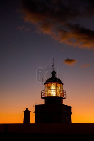 Foto de Faro iluminado con una lámpara encendida durante la puesta del sol. Nubes dramáticas en el fondo - Imagen libre de derechos