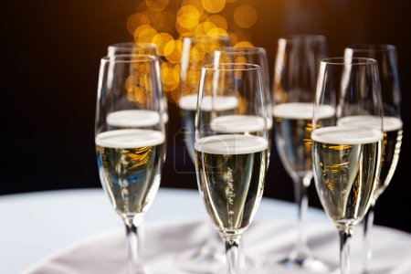 Foto de Elegantes flautas de champán llenas de vino espumoso, bellamente arregladas sobre un paño blanco, con un telón de fondo de luces bokeh doradas creando un ambiente festivo. - Imagen libre de derechos