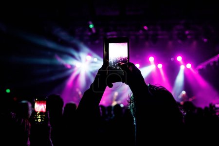 Téléchargez les photos : Une main tient un smartphone, enregistrant un événement animé éclairé par des lumières colorées. - en image libre de droit