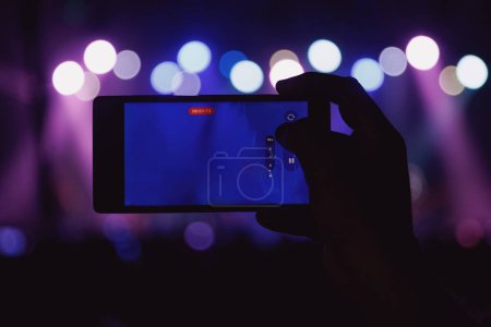 Téléchargez les photos : Une main tient un smartphone, enregistrant un événement animé éclairé par des lumières colorées. - en image libre de droit