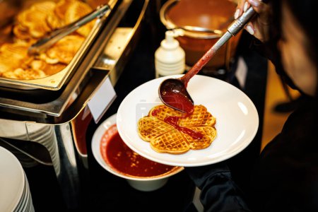 A visitor puts a waffle with berry sauce on a plate during a buffet breakfast at a hotel
