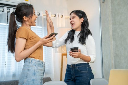 Photo for Two beautiful Asian girls holding smartphones clapping their hands hifive, happy to order food via online app to deliver to work successfully - Royalty Free Image