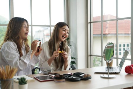 Zwei asiatische Mädchen, Partner und Mitbewohner verkaufen online Kosmetika, führen Puder, Lippenstift, Pinsel ein, benutzen Smartphones und Laptops, um in einer Wohnung zu arbeiten.