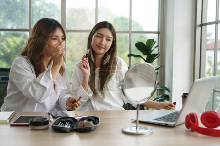 Zwei junge asiatische Blogger, Mitbewohner, die online Kosmetika verkaufen und Puder, Lippenstifte, Pinsel und Make-up-Bewertungen mit Smartphones und Laptops in ihrer Wohnung empfehlen.