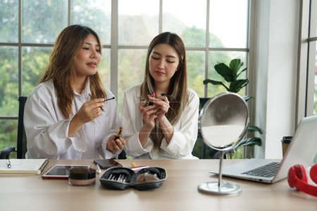 Zwei junge asiatische Blogger, Mitbewohner, die online Kosmetika verkaufen und Puder, Lippenstifte, Pinsel und Make-up-Bewertungen mit Smartphones und Laptops in ihrer Wohnung empfehlen.