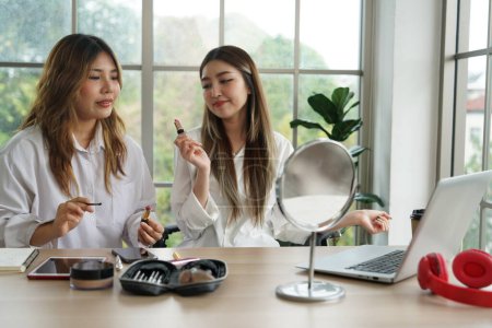 Zwei asiatische Mädchen, Partner und Mitbewohner verkaufen online Kosmetika, führen Puder, Lippenstift, Pinsel ein, benutzen Smartphones und Laptops, um in einer Wohnung zu arbeiten.