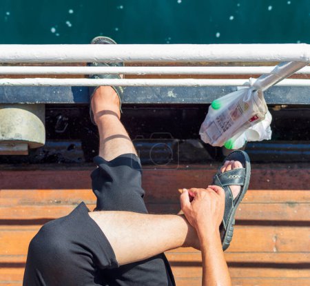Téléchargez les photos : Vue du dessus de la personne aux jambes croisées, assise sur un étroit passage maritime d'un traversier, regardant vers la mer, avec un sac en plastique suspendu à la rampe de passage contenant un journal et des bouteilles d'eau - en image libre de droit