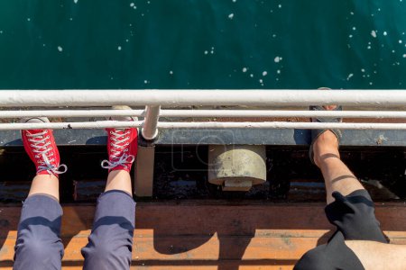 Téléchargez les photos : Vue de dessus montrant les jambes de deux personnes, assises sur un étroit passage côté mer d'un traversier, regardant vers la mer, et mettant leurs jambes sur la main courante de passage - en image libre de droit