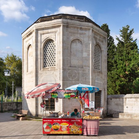 Foto de Istanbul, Turkey - September 1, 2022: Young man selling corn, potatoes and ice cream on traditional Turkish fast food cart with historical tomb in the background at Eyupsultan district - Imagen libre de derechos