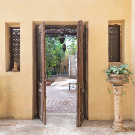 Téléchargez les photos : Porte ouverte décorée en bois antique servant de médiateur à deux fenêtres en bois et menant à une cour paisible avec verdure luxuriante et escalier en pierre - en image libre de droit