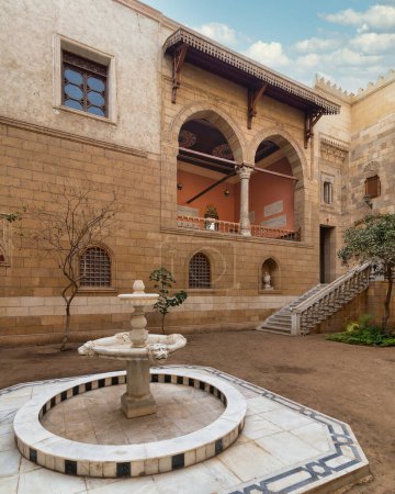 Serene courtyard showcases the timeless beauty of Mamluk architecture with high big arches and stonework with welcoming staircase leading to ornate balcony. A classic marble fountain sits at center