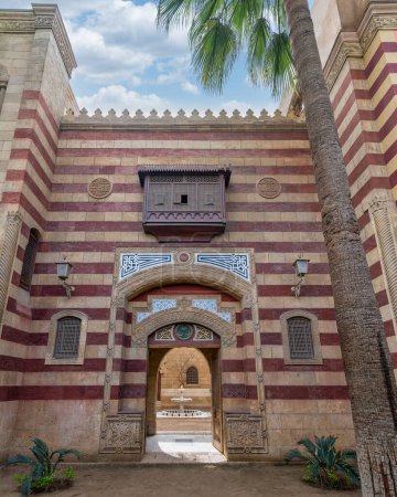 Auffällige rot-weiß gestreifte bogenförmige Eingangstür führt in ein Gebäude im Stil der Mamluk-Ära, in dem komplexe architektonische Details von üppigen Palmen umgeben sind