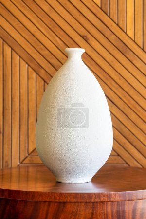Un jarrón blanco texturizado se apoya sobre una mesa de madera pulida, complementada por las diagonales de un revestimiento de madera