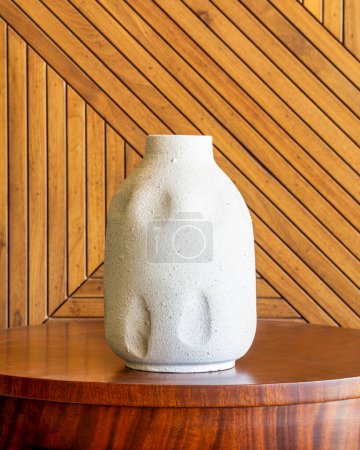 Un jarrón blanco texturizado se apoya sobre una mesa de madera pulida, complementada por las diagonales de un revestimiento de madera