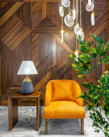 Sillón naranja moderno, pantalla de lámpara en una pequeña mesa de madera, macetero con arbustos verdes y candelabro de cristal alto contemporáneo, en un pasillo con pared de revestimiento de madera decorada y suelo de mármol blanco