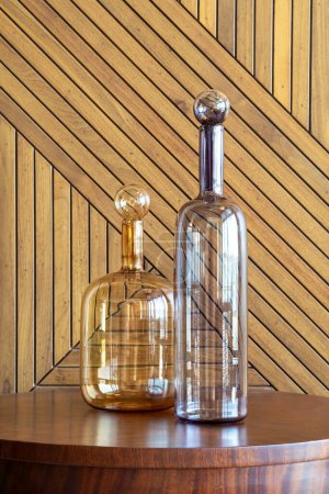 Deux carafes en verre transparent avec bouchons ornés reposant sur une surface en bois poli, complétées par une toile de fond en bois motif chevrons