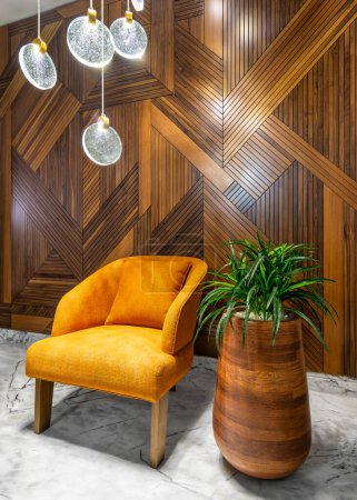 Sillón naranja moderno, junto a una maceta de madera alta con arbustos verdes y una lámpara de araña de cristal alta contemporánea, en un pasillo con pared de revestimiento de madera decorada y suelo de mármol blanco