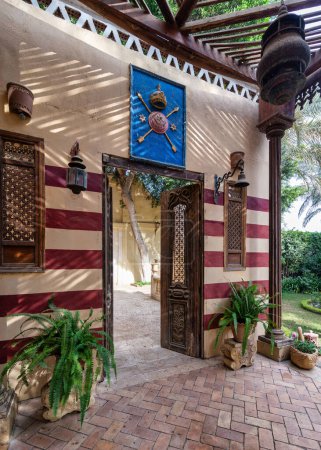 Une porte en bois magnifiquement conçue s'ouvre sur une cour avec un mur à rayures rouges et blanches, des colonnes en bois et des pots de plantes verdoyantes