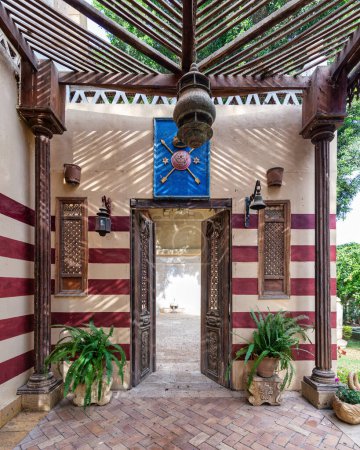 Una puerta de madera bellamente hecha a mano se abre a un patio con paredes de rayas rojas y blancas, columnas de madera y exuberantes macetas de plantas verdes.