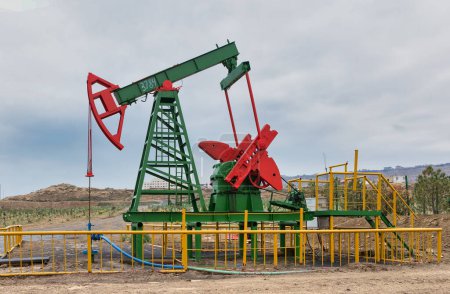 An operational green and red oil pumpjack surrounded by yellow fencing, set against a cloudy sky, in Baku, Azerbaijan