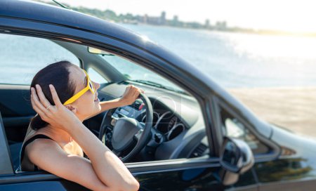 Téléchargez les photos : Belle jeune femme dans une voiture souriant dans des lunettes de soleil jaunes sur fond de mer. - en image libre de droit