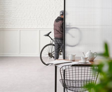 Téléchargez les photos : Homme avec vélo dans la chambre, table de chaise bureau brique blanche et mur classique, feuille vert flou. - en image libre de droit