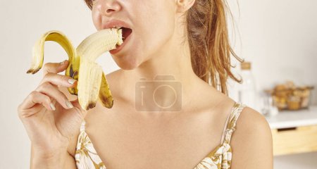 Foto de Mujer comiendo plátano, primer plano, mano y boca. Fondo de cocina. - Imagen libre de derechos