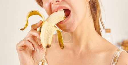 Téléchargez les photos : Femme mangeant de la banane, gros plan, main et bouche. Fond de cuisine. - en image libre de droit