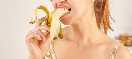 Photo for Woman eating banana, close-up, hand and mouth. Kitchen background. - Royalty Free Image