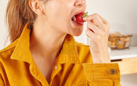Téléchargez les photos : Femme mangeant des fraises, gros plan, la main et la bouche. Fond de cuisine. - en image libre de droit