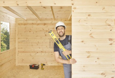 Foto de Construction man, builder man construct new wooden home, tiny house style. Spirit level, screw, tool bag and charged drill. Wood shelter style. - Imagen libre de derechos