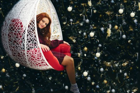Téléchargez les photos : Jeune jolie femme dans une chaise berçante en osier près de l'arbre du Nouvel An - en image libre de droit