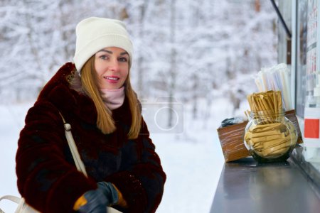 Téléchargez les photos : Jolie femme achetant une boisson chaude à un kiosque dans un parc d'hiver, arbres enneigés - en image libre de droit