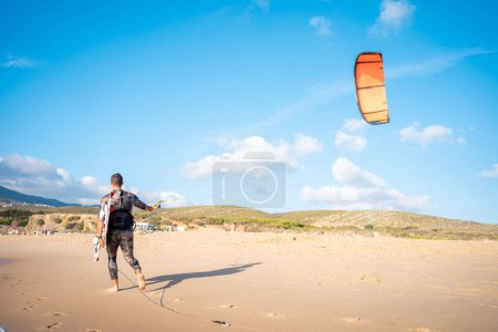 Portrait vague kitesurfer marchant au vent à la plage avec sa planche et cerf-volant. Homme kite surfer promenade plage de sable océan avec sa planche de kite surf.