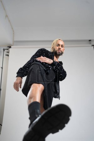 Full length gay man wearing black skirt and big black boots doing step on camera. Afro-american homosexual posing in photo studio on white background. Bearded gay with make up. Photo studio