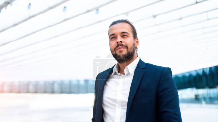 Successful businessman in suit with beard standing in front of office building confidently looking away. Hispanic male business person side view portrait. Web banner, Free space